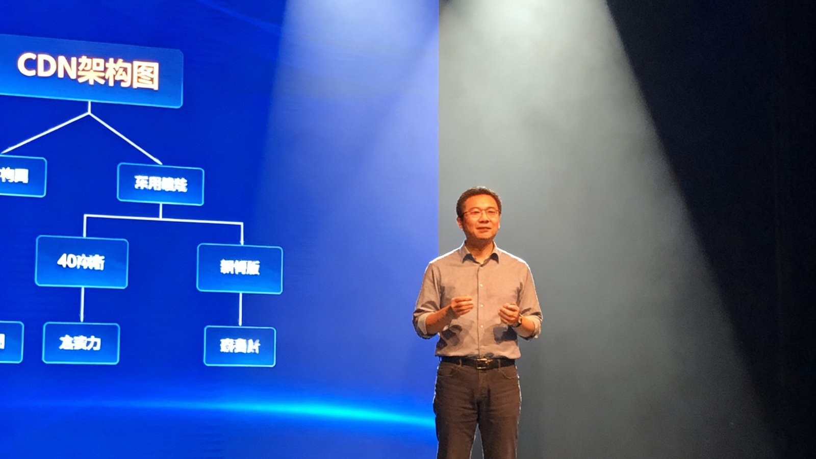 一位40岁左右的技术专家站在舞台中央演讲，背景大屏幕显示着CDN架构图，灯光聚焦在他身上